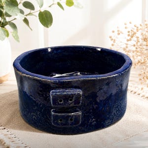 May include: A handcrafted, dark blue ceramic bowl with a textured surface. The bowl has a wide, open design with two rectangular, decorative elements on the side. The bowl is sitting on a beige linen surface.