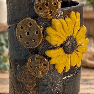 May include: A cylindrical ceramic vase with a dark, textured, charcoal-colored glaze. The surface features embossed floral patterns, four small circular brown discs with holes, and a prominent, three-dimensional yellow sunflower sculpture.
