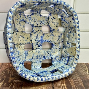 May include: A ceramic basket with a woven design. The basket is off-white with blue speckles and a blue rim. The basket is sitting on a wooden surface.