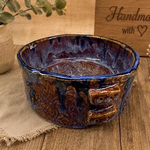 May include: A handmade ceramic bowl with a unique glaze. The bowl features a deep blue rim and exterior with brown and blue mottled patterns. Two small handles are on the side. The bowl is on a wooden surface.
