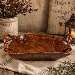 May include: A rectangular brown ceramic serving dish with two curved handles on opposite sides. The dish features a glossy, textured finish with subtle embossed patterns along the edges and a deep, rich brown glaze throughout the interior and exterior.