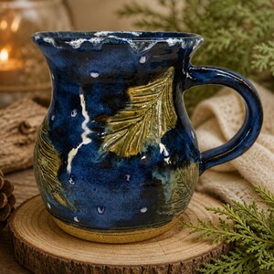 May include: A handcrafted ceramic mug with a dark blue glaze and leaf design. The mug features a rounded body, a sturdy handle, and is approximately 4 inches tall. The design includes white speckles and a light brown base, perfect for enjoying hot beverages.