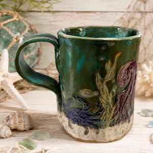 May include: A handmade ceramic mug with a glossy teal and green glaze featuring raised relief designs of a dark blue crab, a purple octopus, and seaweed. The base of the mug has a textured, sandy-colored finish, and it includes a curved handle.