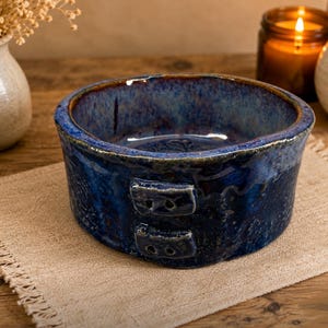 May include: A handmade, round, blue ceramic bowl with a glossy finish. The bowl has a textured surface and two rectangular handles on the side. The rim is a darker shade of blue. The bowl is sitting on a tan linen placemat.