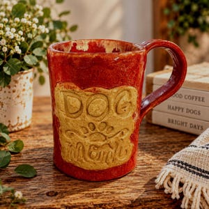 May include: A glossy red ceramic mug with a large, textured tan rectangular patch on the front. The patch features the embossed text "DOG MOM" with a paw print icon centered between the words. The mug has a curved handle and a smooth finish.
