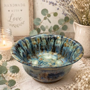 May include: A ceramic bowl featuring a reactive glaze with swirling patterns of deep blue, teal, and earthy brown tones. The bowl has a wide, flared rim and a rounded base, showcasing a glossy finish with visible color variations throughout the surface.