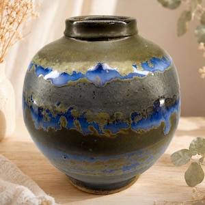 May include: A handcrafted ceramic vase with a rounded body and a dark, textured finish. The vase features bands of blue and green glaze, creating a unique, artistic design. The vase is a decorative item.
