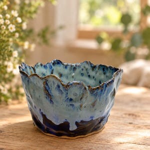 May include: A handmade ceramic bowl with an irregular, wavy rim. The exterior features a deep navy blue base that transitions into a lighter, mottled sky blue glaze. The surface has a textured, organic finish with visible indentations.