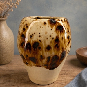 May include: A ceramic tumbler with a speckled brown and cream glaze. The handmade pottery has a rounded shape with a lighter base and a darker, textured top. The cup is suitable for beverages.