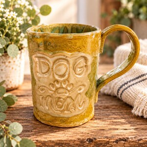 May include: A handmade ceramic mug with a glossy mustard yellow and green glaze. The front features a raised, cream-colored rectangular panel embossed with the text DOG MOM and a paw print graphic. It has a sturdy, curved handle on the right side.