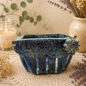 May include: A rectangular ceramic berry basket with a glossy blue and dark teal glaze. The exterior features a honeycomb texture, vertical cutouts, and the embossed text "BEEYOU..." with a small sculpted flower attached to the top right corner.