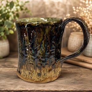 May include: A ceramic mug with a textured, hand-thrown appearance, featuring a glossy glaze that transitions from a deep, dark forest green and black at the top to a lighter, sandy beige at the base, with a relief pattern of pine trees.