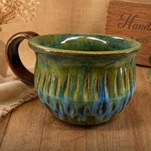 May include: A handmade ceramic mug with a textured, carved design. The mug features a green and blue glaze with a brown handle. The mug is on a wooden surface. The mug is a unique piece of pottery.