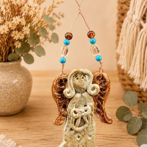 May include: A ceramic angel ornament with a speckled cream finish and brown glazed wings. The figure holds a small circular object and hangs from a copper wire loop adorned with two amber, two clear, and four bright turquoise glass beads.
