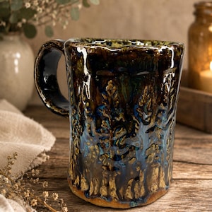 May include: A handmade ceramic mug with a glossy, textured glaze in deep shades of forest green, dark blue, and earthy brown. The surface features a raised, embossed pattern of pine trees wrapping around the exterior, with a sturdy, curved handle.