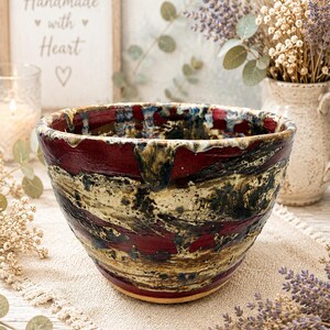 May include: A ceramic bowl featuring a deep red, black, and cream-colored glaze with a textured, dripped finish along the rim. The bowl has a wide, rounded shape with a tapered base, showcasing a complex, layered pattern of dark and light colors.