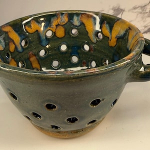 Può includere: Colino in ceramica fatto a mano, esterno verde scuro e interno maculato con accenti arancioni e blu. Il colino ha un manico e fori di drenaggio.