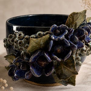 May include: A dark blue ceramic bowl with a floral design. The bowl is adorned with sculpted blue flowers, green leaves, and textured accents. The handmade pottery piece is suitable for home decor.