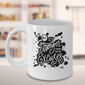 Könnte beinhalten: Weiße Keramik-Kaffeetasse mit schwarzem Spritzmuster und dem Text "Sei stärker als deine Ausreden".