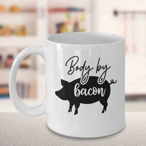 Puede incluir: Taza de café de cerámica blanca con una silueta negra de un cerdo y el texto "Body by bacon".