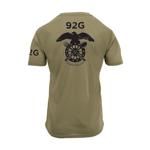 May include: Olive green t-shirt with black graphic on the back. The graphic includes an eagle above a fork, spoon, and wheel design, with the text "92G" and "Culinary Specialist". The left sleeve has the text "2G".