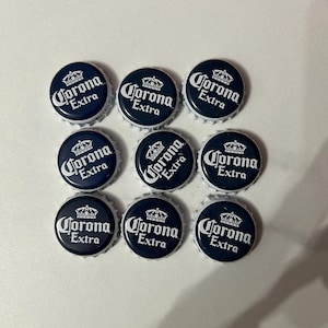May include: Nine Corona Extra bottle caps arranged in a grid. Each cap is dark blue with white text and a crown logo. The caps have a silver, crimped edge.