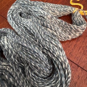 Puede incluir: Una madeja de hilo que presenta una mezcla jaspeada de fibras gris oscuro, gris claro y blanco. El hilo está retorcido en una bobina suelta y asegurado con un lazo de cuerda amarillo brillante, mostrando una apariencia texturizada y multitonal.