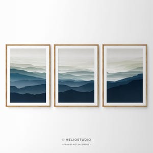 Puede incluir: Conjunto de tres impresiones abstractas de paisajes con una cadena montañosa en capas en tonos azules y grises. Las impresiones están enmarcadas en madera natural.