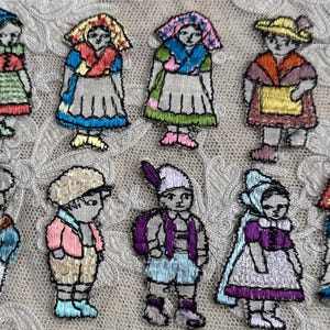 May include: A collection of ten embroidered figures of children in traditional clothing. Each figure is approximately 2 inches tall and features detailed stitching in various colors, including blue, red, yellow, and green. The figures are arranged on a lace background.