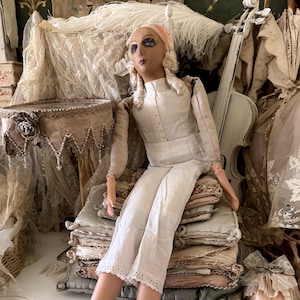 May include: A vintage doll with blue eyes, wearing a white outfit and gold heels, sits atop a stack of pillows. The doll has a pink headscarf and is surrounded by antique decor, including a white violin and lace accents.