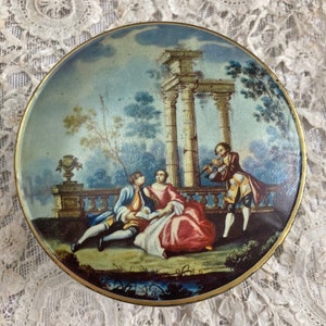 May include: A round, metal trinket box featuring a painted scene of four figures in a garden setting. The scene depicts a couple seated on a stone ledge, a woman standing behind them, and a man playing a musical instrument. The background includes a stone archway, trees, and a blue sky.