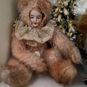 May include: A vintage doll with a porcelain face and a pink fur body. The doll is wearing a pink fur hood and a ruffled collar. The doll is sitting on a white shelf. The background includes a floral arrangement.
