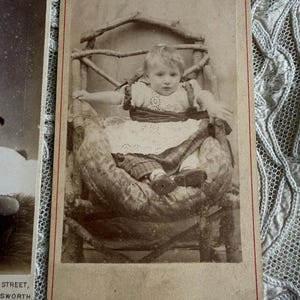 May include: Sepia-toned antique photograph of a young child seated in a rustic wooden chair. The child is wearing a white dress with a dark ribbon and is surrounded by a patterned blanket. The photograph has a red border.