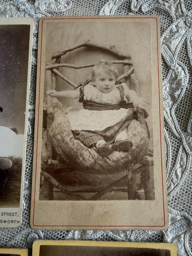 May include: Sepia-toned antique photograph of a young child seated in a rustic wooden chair. The child is wearing a white dress with a dark ribbon and is surrounded by a patterned blanket. The photograph has a red border.