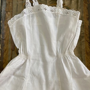 May include: A white cotton slip dress with a scalloped lace trim and adjustable straps. The dress has a gathered waist and a tiered skirt.