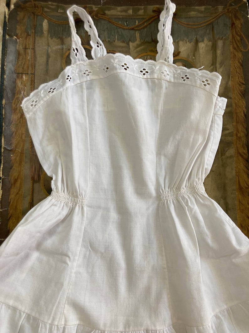 May include: A white cotton slip dress with a scalloped lace trim and adjustable straps. The dress has a gathered waist and a tiered skirt.