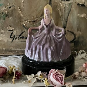 Könnte beinhalten: Eine Porzellanfigur einer Frau in einem Lavendelkleid mit schwarzem Sockel. Die Figur steht auf einer weißen Oberfläche mit rosa Blumen.