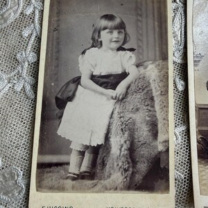 May include: Vintage black and white photograph of a young girl in a white dress with a dark sash, standing next to a textured surface. The photograph is labeled "F. HIGGINS, HOLYROOD ST, CHARD" at the bottom.