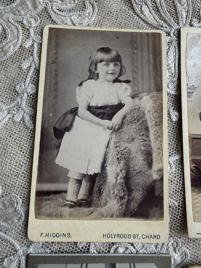 May include: Vintage black and white photograph of a young girl in a white dress with a dark sash, standing next to a textured surface. The photograph is labeled "F. HIGGINS, HOLYROOD ST, CHARD" at the bottom.