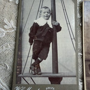 May include: Vintage black and white photograph of a young child in a dark suit with a white lace collar, standing on a platform. The photograph is mounted on a gray card with the text "W.C. Murphy, Prize Medallist, Bideford."
