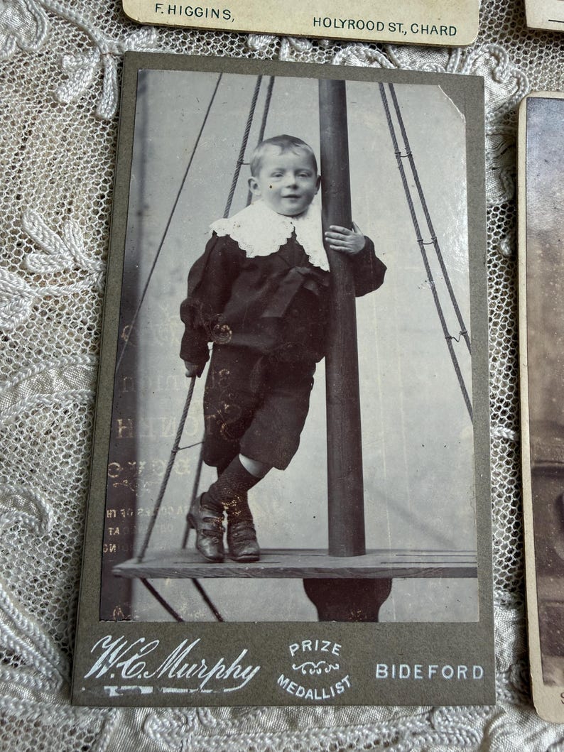 May include: Vintage black and white photograph of a young child in a dark suit with a white lace collar, standing on a platform. The photograph is mounted on a gray card with the text "W.C. Murphy, Prize Medallist, Bideford."