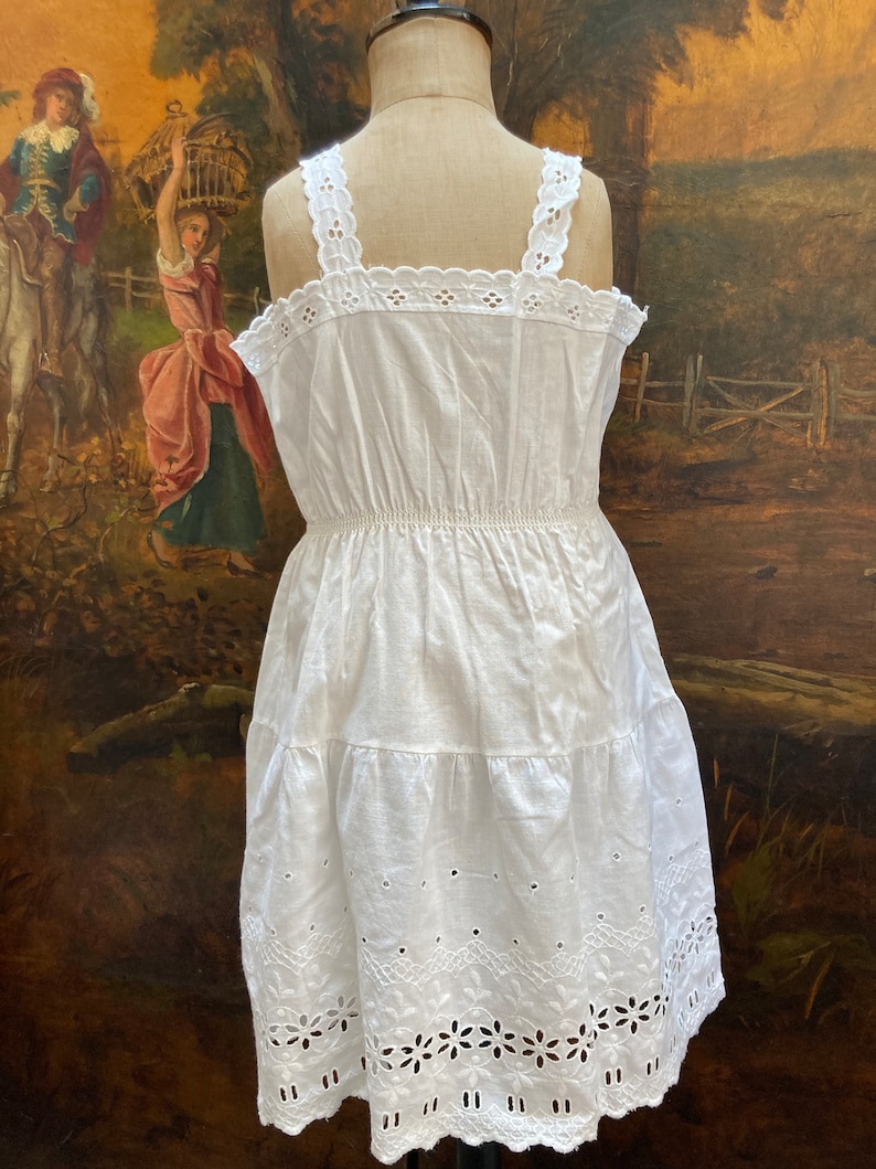 May include: White cotton dress with eyelet lace trim and a tiered skirt. The dress has a fitted bodice and a gathered waist.