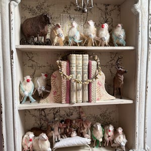 Puede incluir: Un armario blanco envejecido con múltiples estantes, lleno de figuras decorativas de ovejas, libros y una pequeña lámpara de araña. Las ovejas son de varios tamaños y colores, algunas con campanas y cintas. El armario tiene un papel pintado floral.