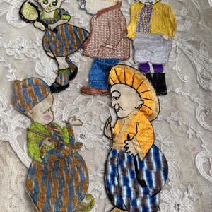 May include: A collection of vintage embroidered fabric figures. The figures depict people in various outfits, including dresses, jackets, and hats. The figures are in shades of blue, brown, yellow, and green, with intricate details.