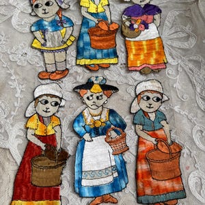 May include: Embroidered patches featuring figures in traditional attire, each holding baskets. The figures wear bonnets and dresses in various colors, including blue, yellow, orange, and red. The patches are likely vintage or handmade.