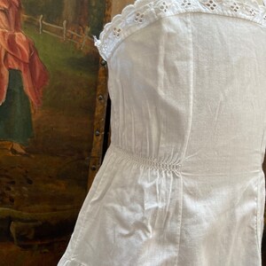 May include: White cotton slip dress with a lace trim around the neckline and straps. The dress has a gathered waist and a tiered skirt.