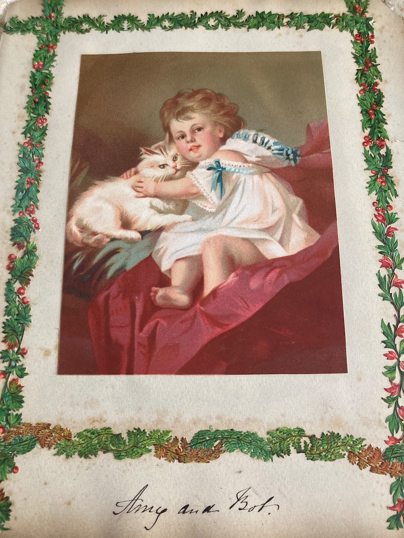 May include: A vintage Christmas card featuring a young girl in a white dress holding a white cat. The card has a green holly border and the text "Amy and Bot." is written in black ink.