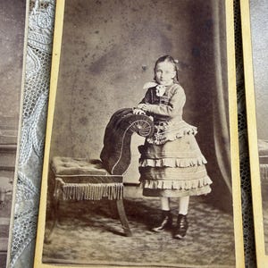 May include: A sepia-toned photograph of a young person in a dress, standing next to a small upholstered bench. The dress has multiple ruffles and the bench has a fringed edge. The photograph is mounted on a card with a yellowed border.