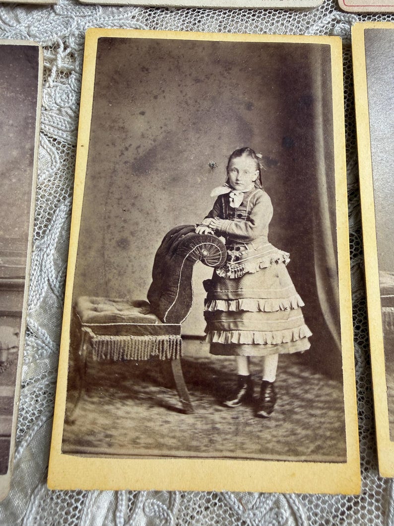May include: A sepia-toned photograph of a young person in a dress, standing next to a small upholstered bench. The dress has multiple ruffles and the bench has a fringed edge. The photograph is mounted on a card with a yellowed border.