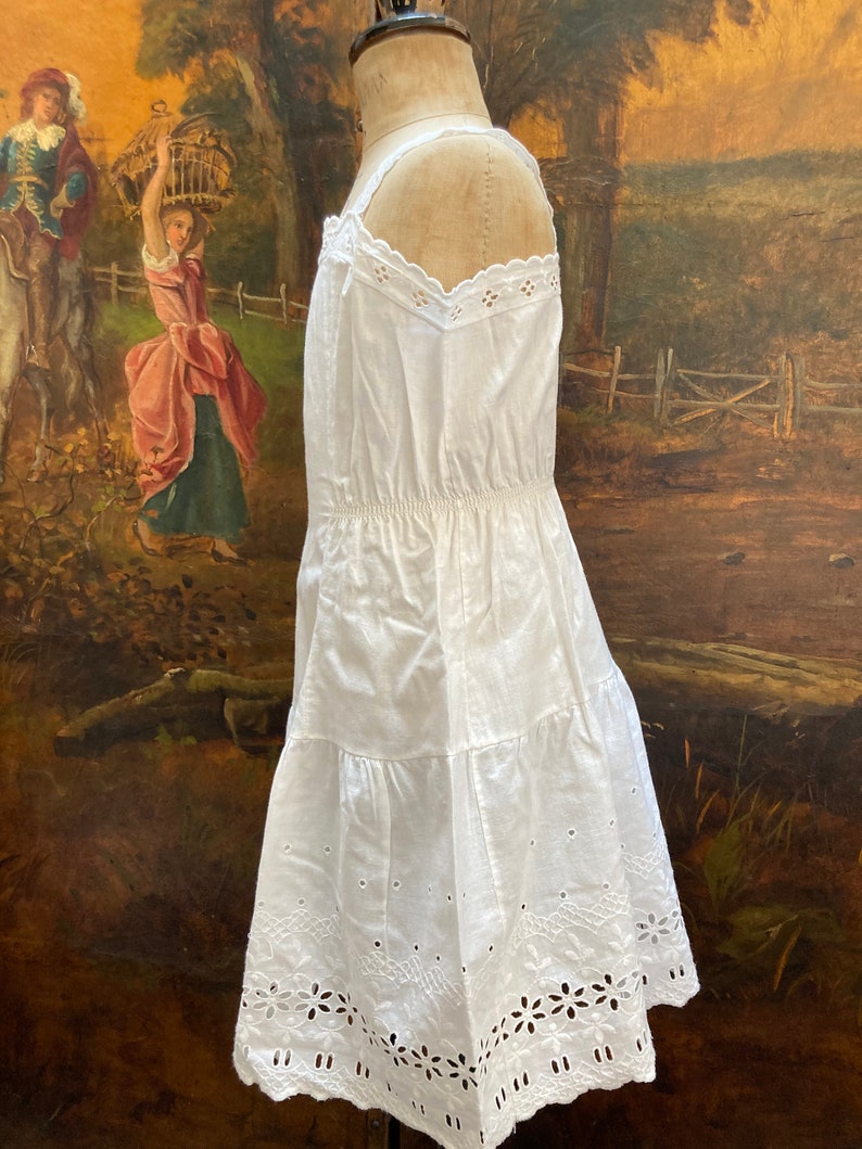 May include: White cotton slip dress with eyelet lace trim. The dress has a gathered waist and a tiered skirt.